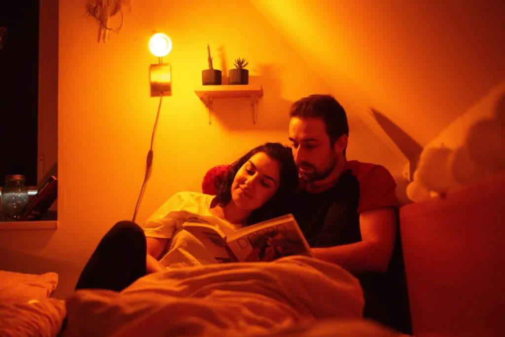 Nástěnná lampa nad nočním stolkem v ložnici a pod ní si pár čte knihu