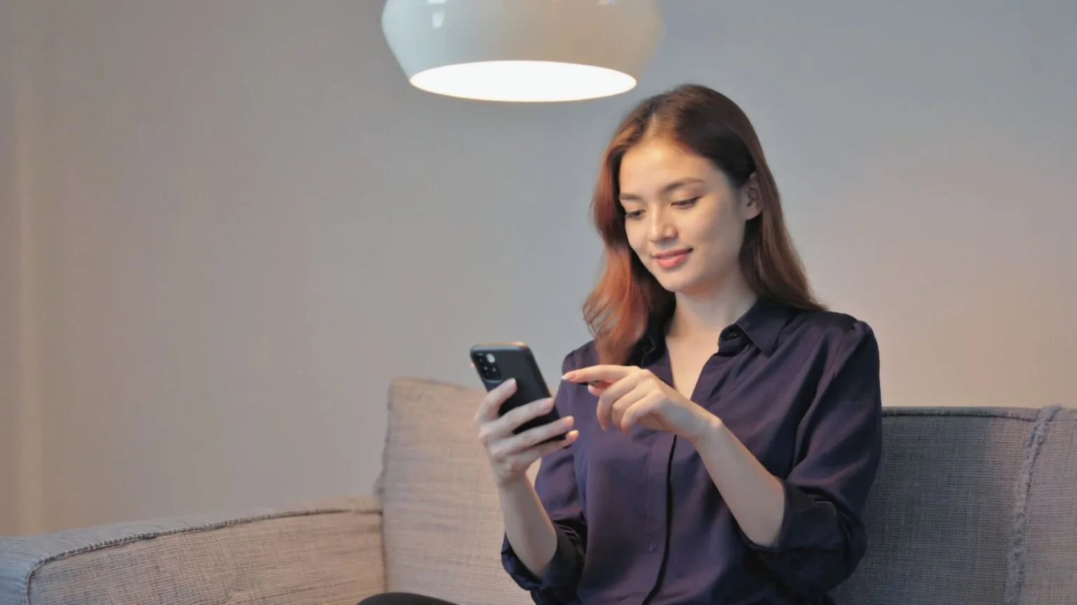 Žena sedí na pohovce a kouká do telefonu a nad ní svítí světlo